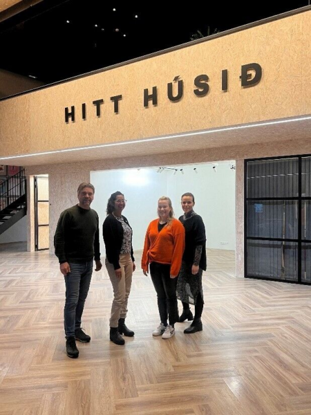 Four people standing in front of a sign saying Hitt Húsið (The Other House, in Icelandic), in a room with wooden floors and wooden walls 