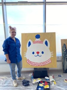 Young person standing in front of an illustration of a cat face in a clown costume and hat, representing that self-care sometimes takes the form of doing what we really want to do and drawing the artwork we like to draw, not what others want us to create