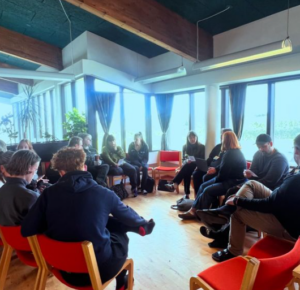 Youth workers sitting in a circle on red chairs, engaging in conversation. Behind them a bright light coming through a window.