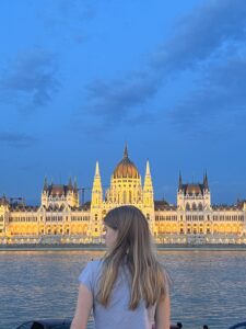 A person with long hair stands by a river, looking at a grand, illuminated building with a dome and spires at dusk. The sky is a deep blue, and the building's lights reflect on the water.