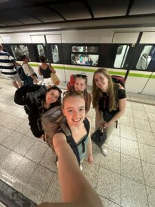 Four young women with backpacks are smiling and taking a selfie on a subway platform. A train is stopped behind them, and other passengers are visible both on the platform and inside the train. The setting suggests they are travelling together.