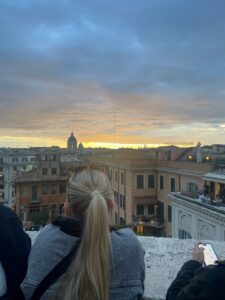  A person with long blonde hair, tied in a ponytail, is looking at a cityscape during sunset. The sky is a mix of blue and orange hues. In the background, a dome of a historic building is visible. Another person is holding a smartphone, possibly taking a photo.